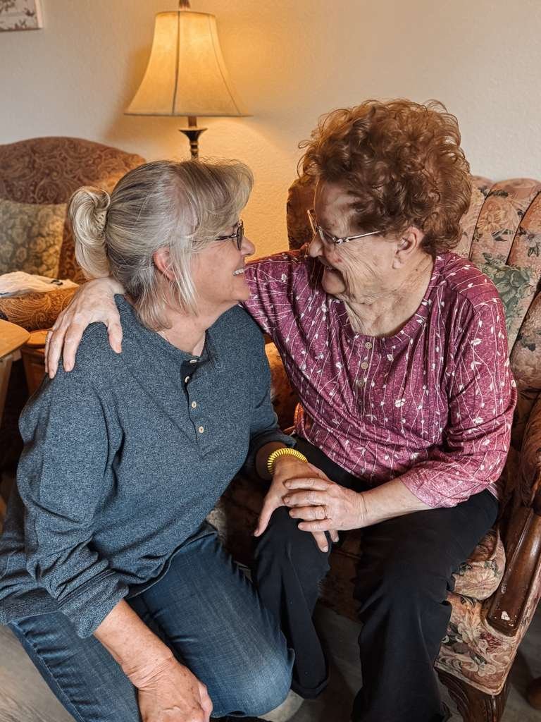 A daughter kneeling beside her mother in an armchair, holding hands and smiling together